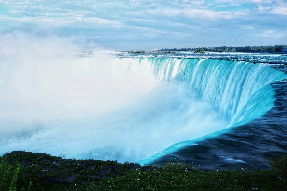 Turquoise water curves over a wide waterfall into rising white mist.