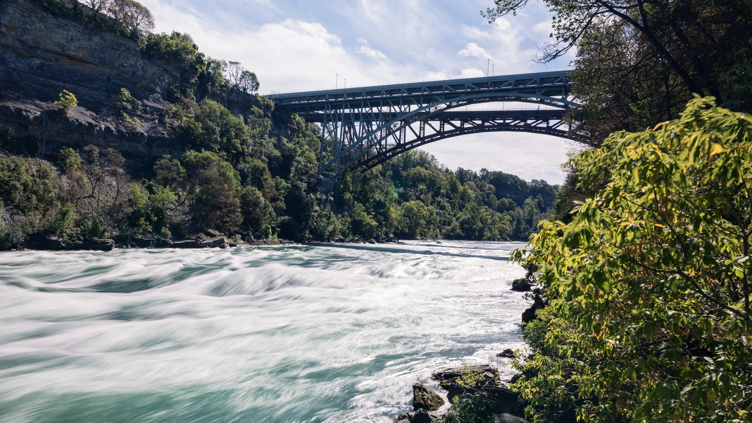 A steel arch bridge spans a rocky gorge above powerful white-water rapids.