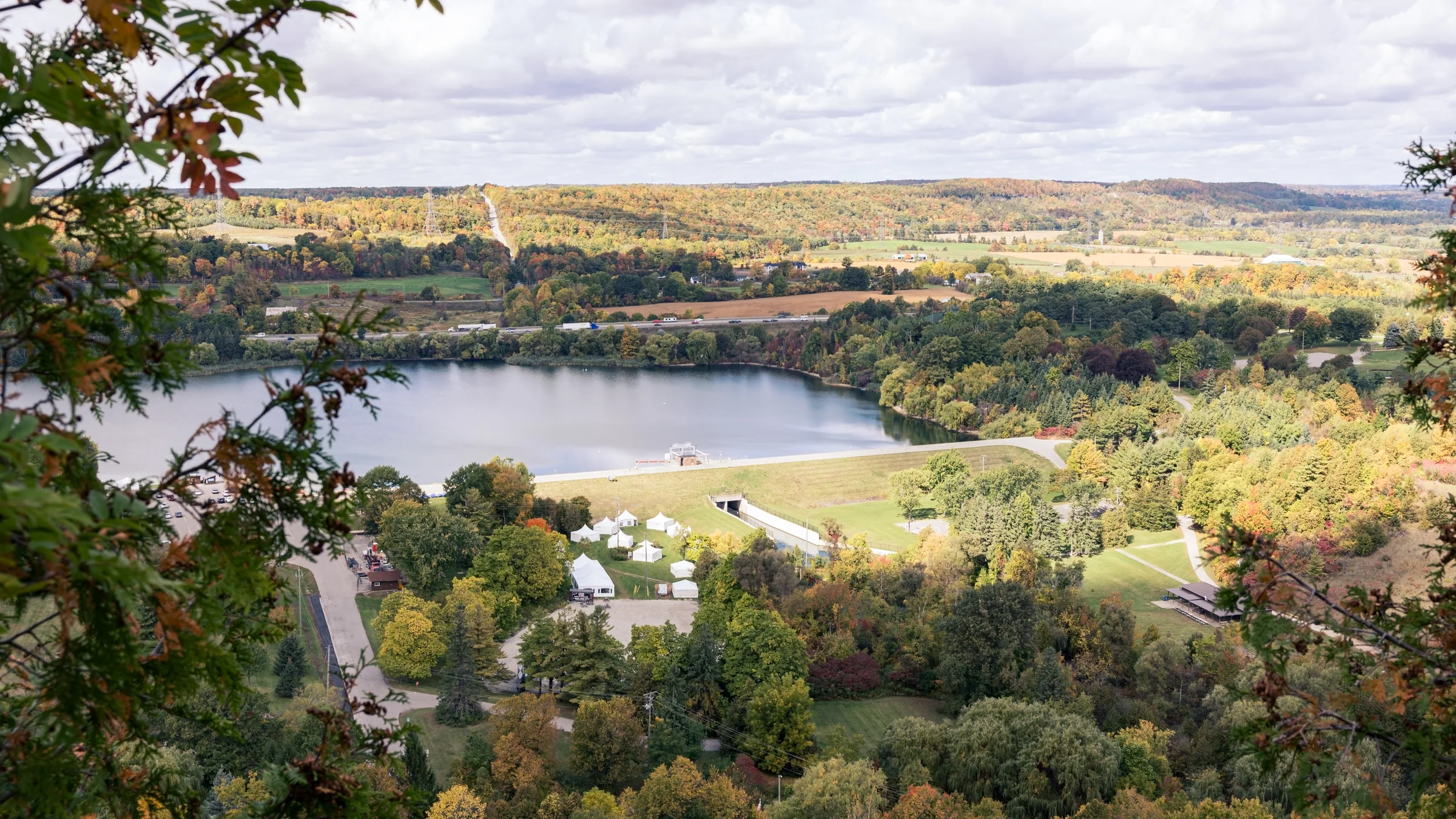 A blue lake and nearby buildings lie among wooded hills turning gold and red.