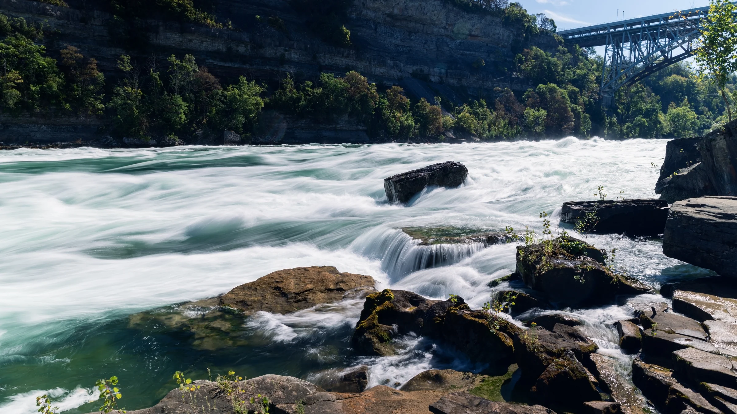 Foaming river water rushes around shoreline rocks below a distant steel bridge.