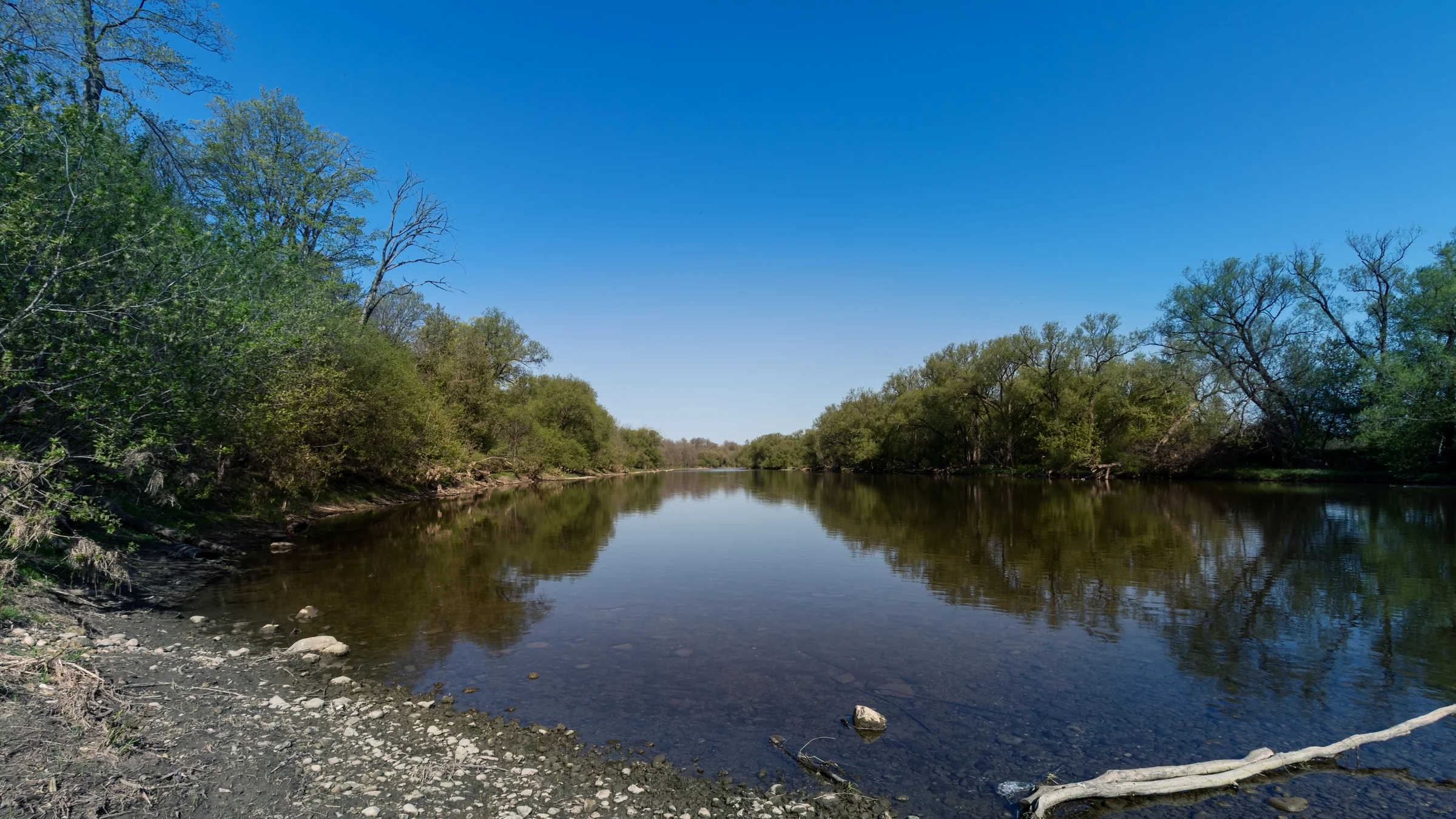A still river reflects a blue sky between two tree-lined banks.