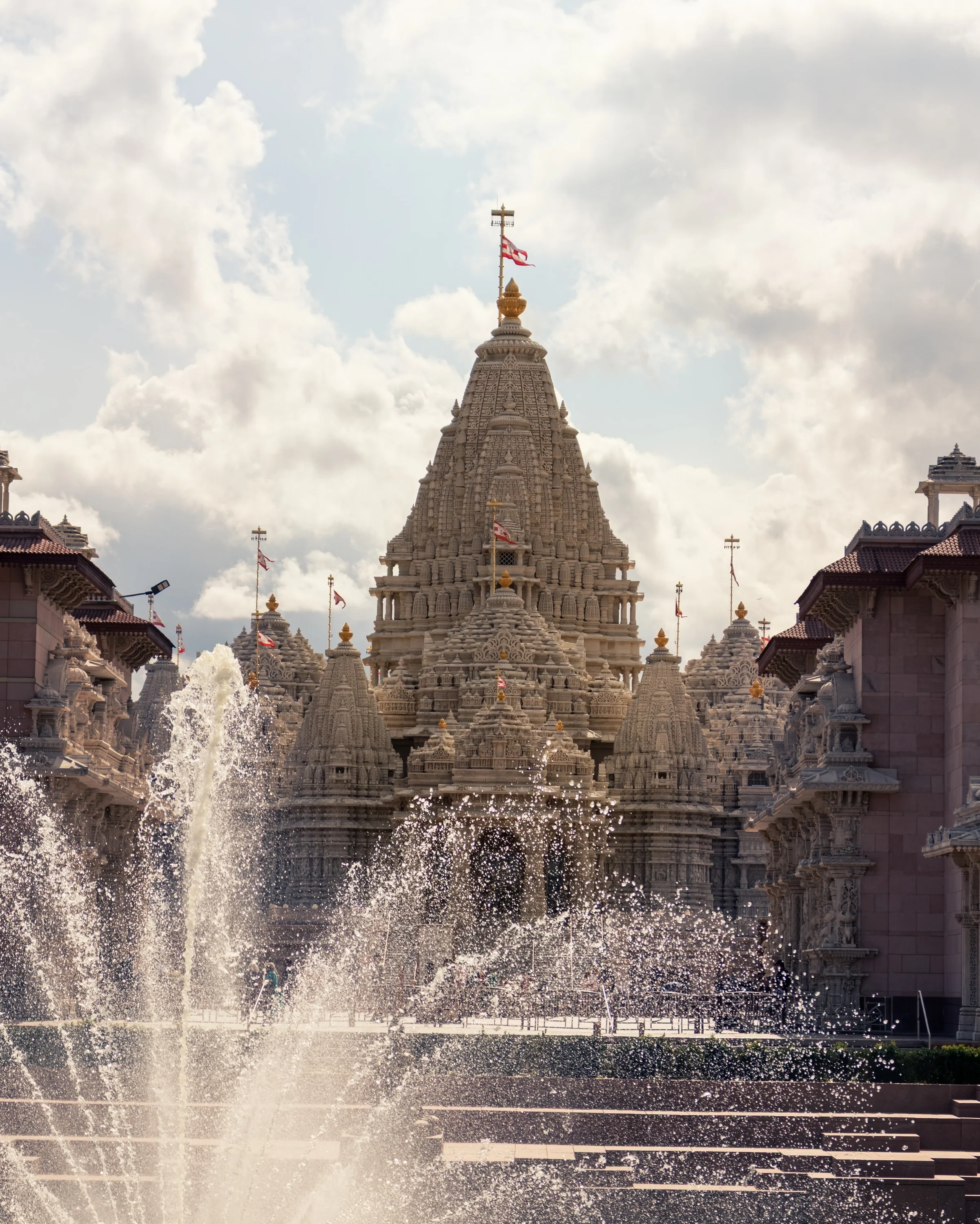 Fountain spray rises in front of an elaborately carved temple beneath a cloudy sky.