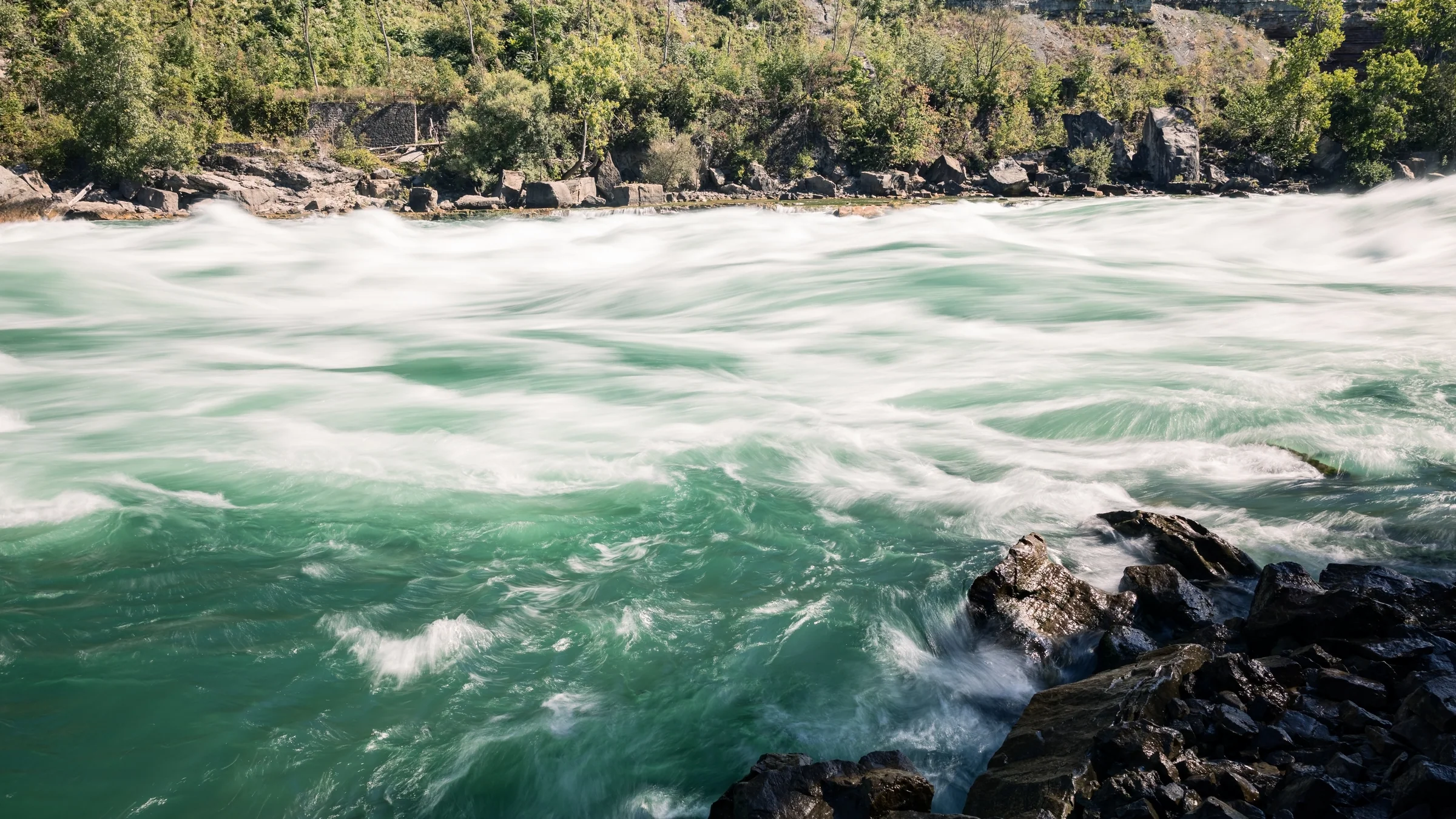 White foam streaks across vivid turquoise water beside dark shoreline rocks.