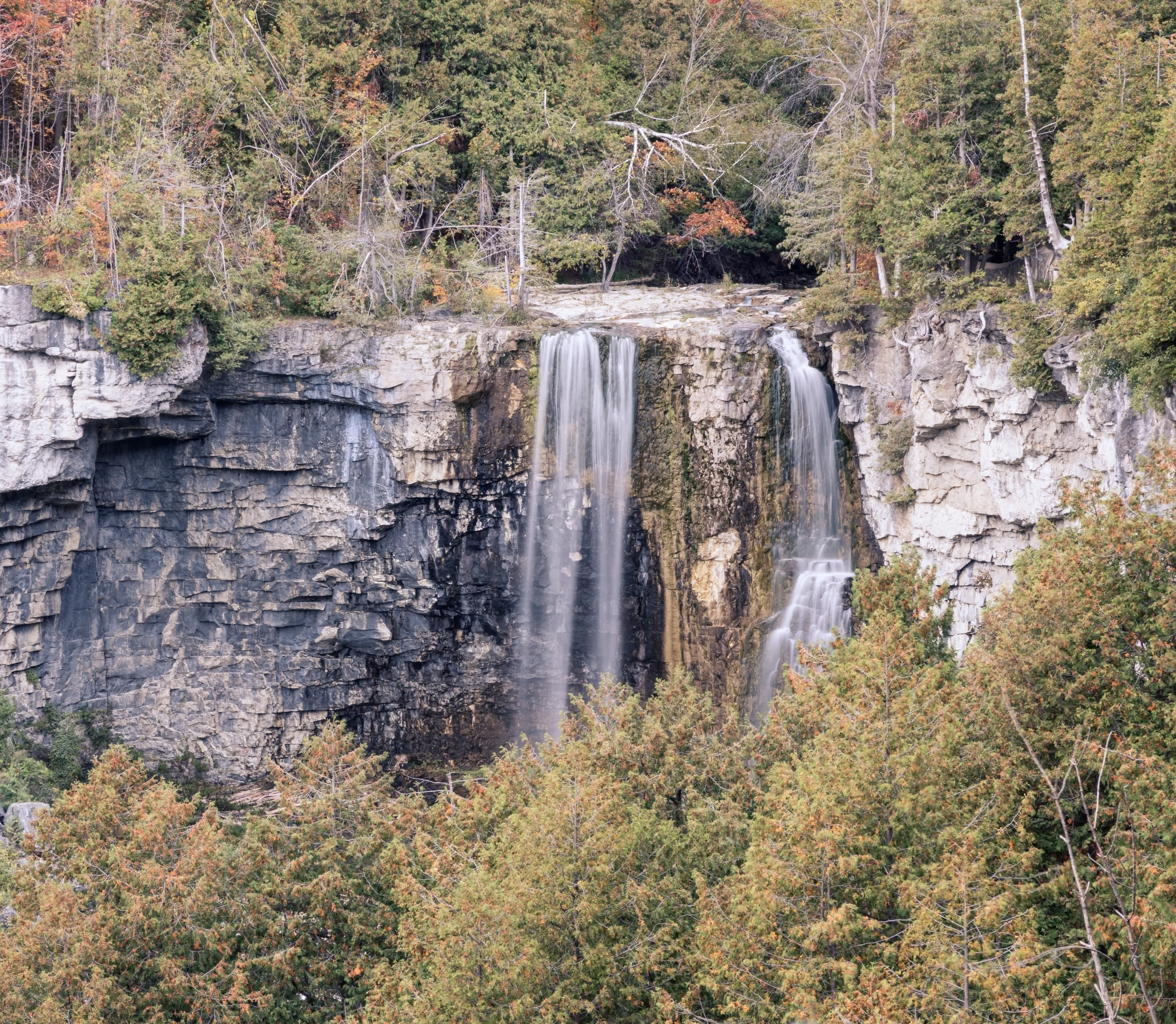 A tall waterfall falls over a layered cliff surrounded by autumn foliage.