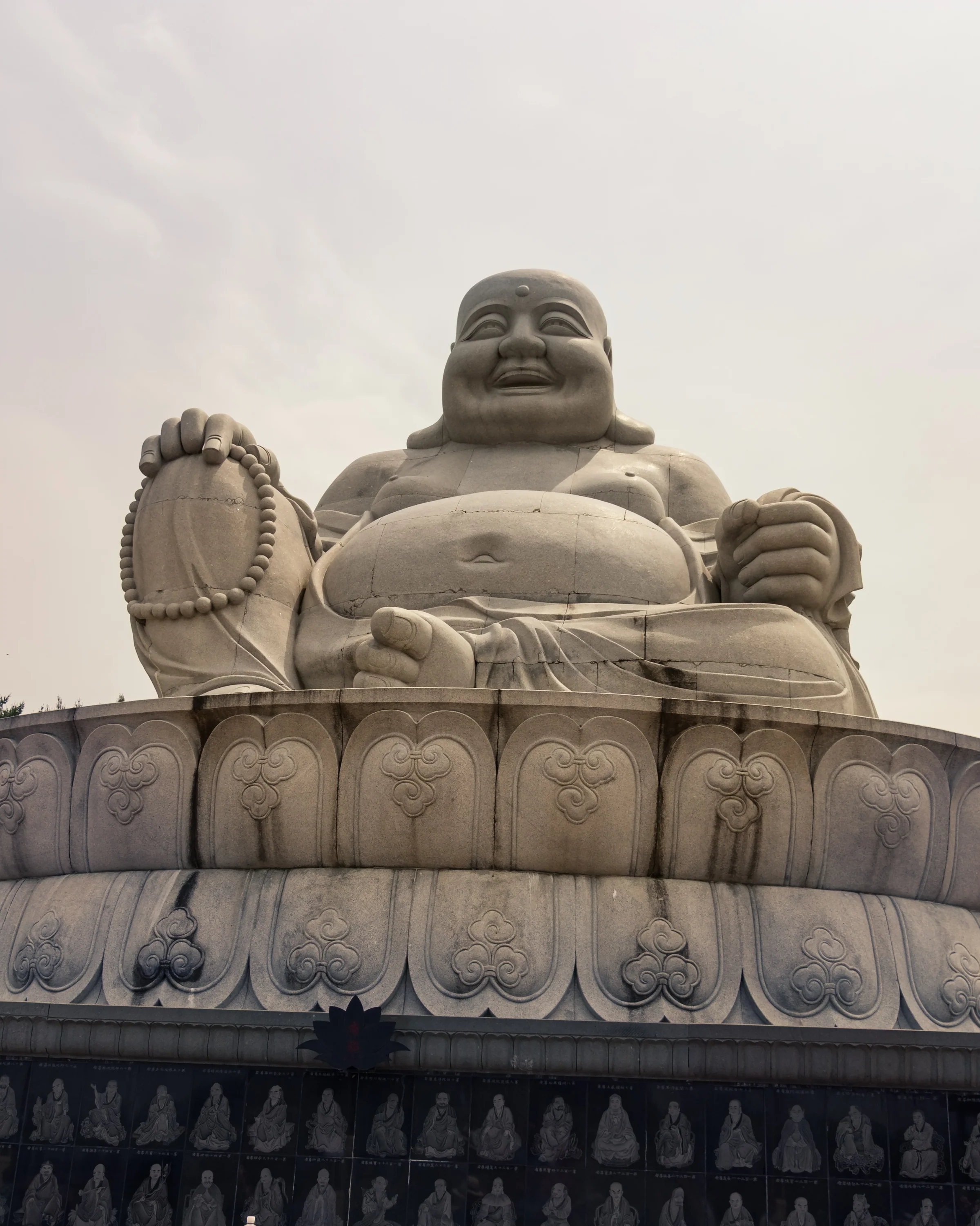 A seated, smiling stone figure rests on a carved pedestal against a pale sky.