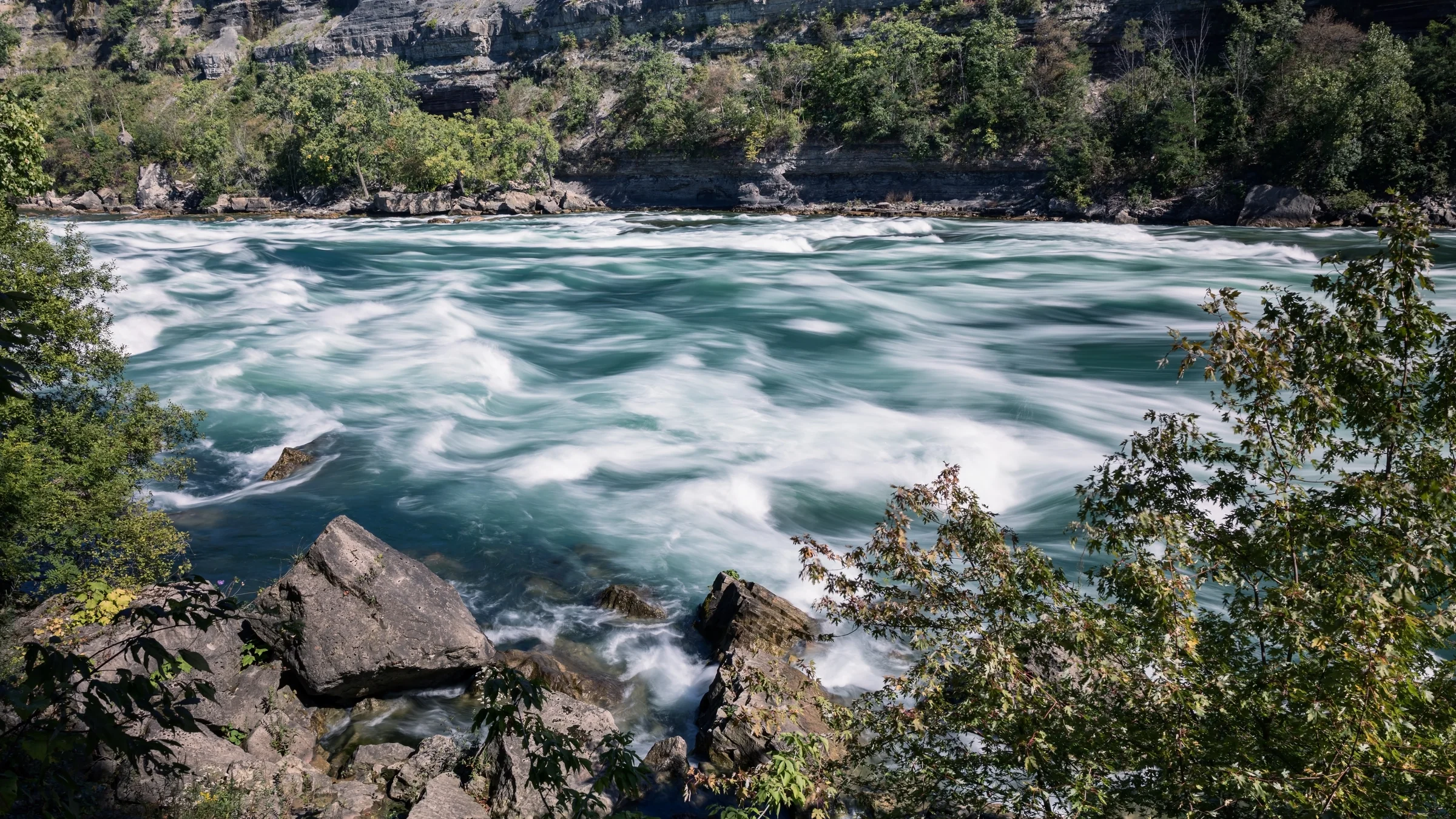 Strong currents form pale swirls across a broad river beneath a wooded cliff.