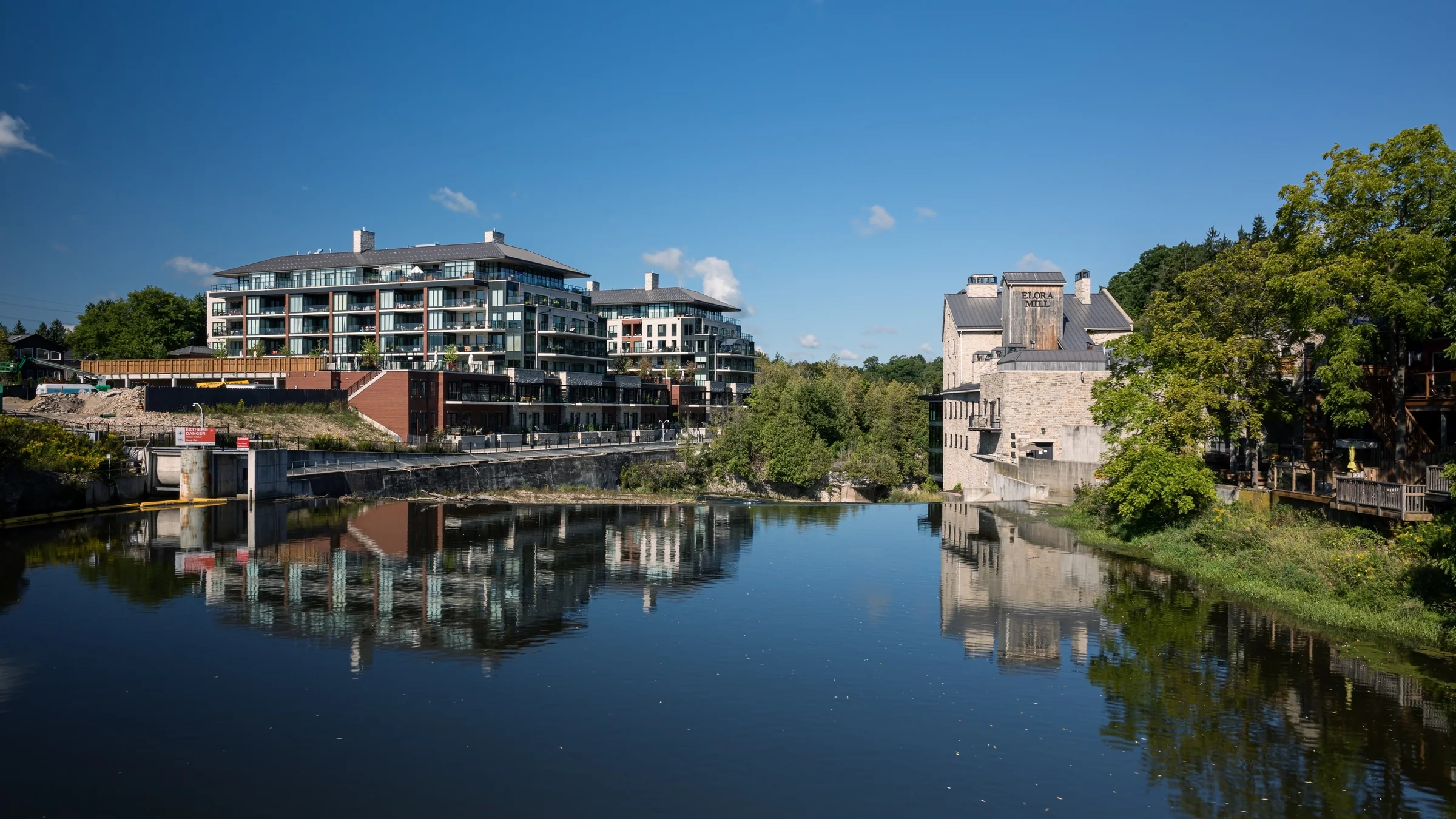 Waterside buildings reflect in a calm pool beside a low dam and rocky riverbank.