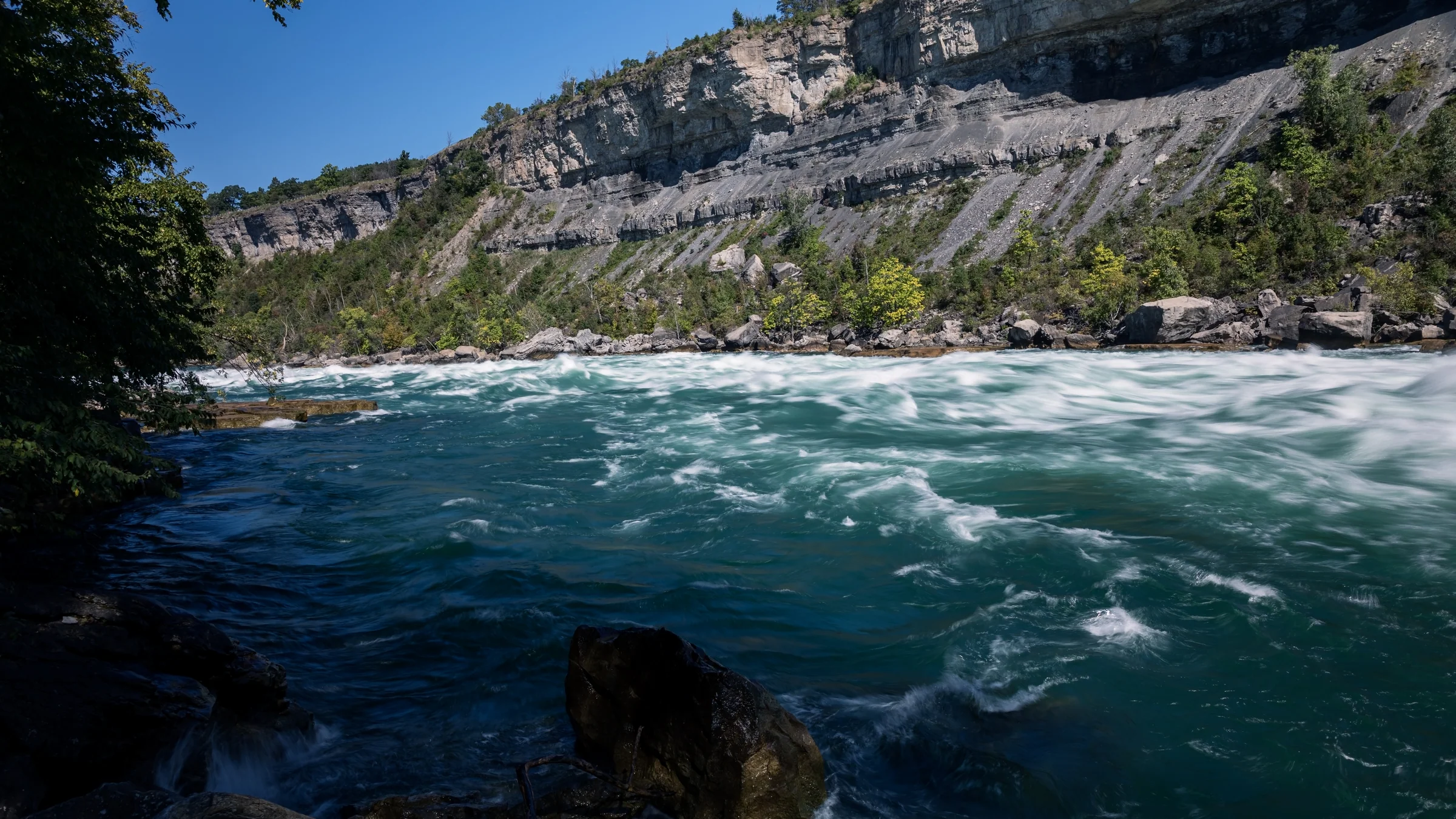 Deep blue and turquoise rapids swirl below a steep layered cliff.