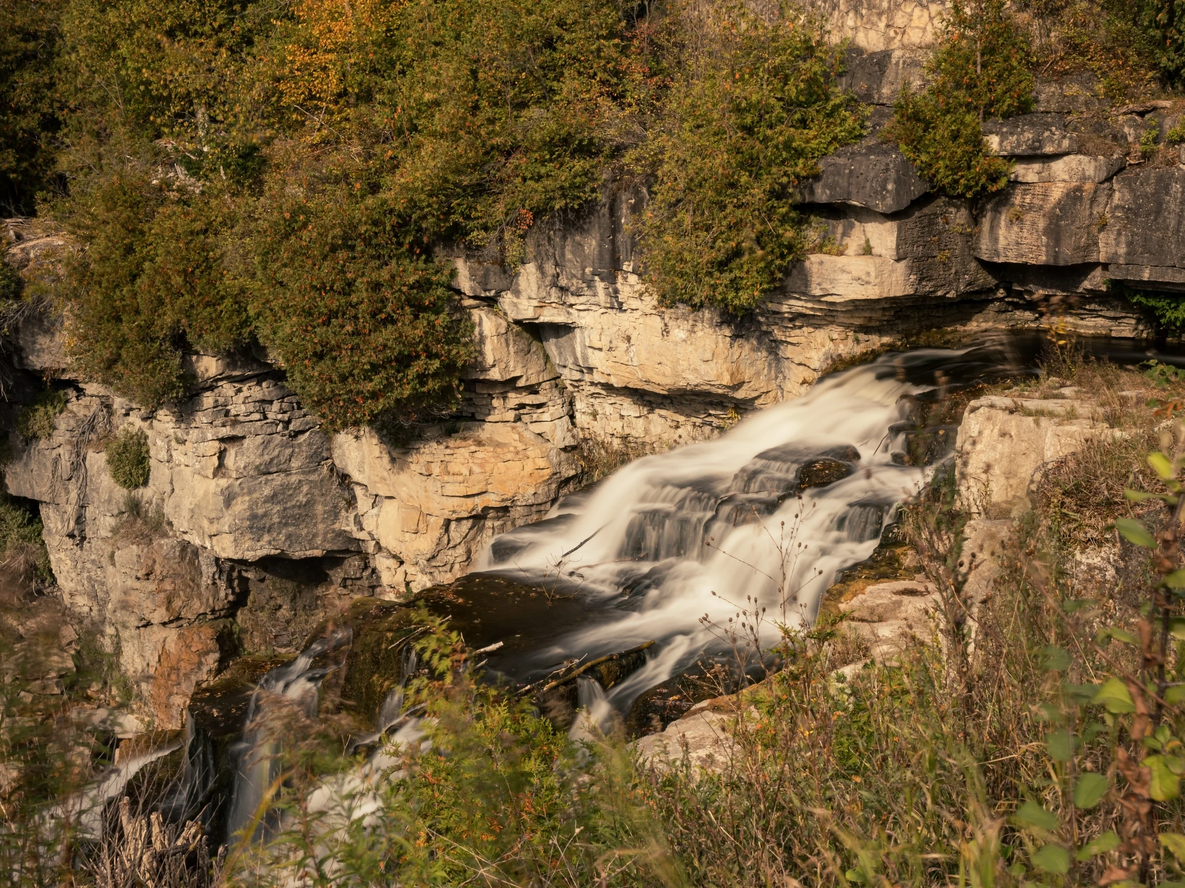 A shallow waterfall slides diagonally across a sunlit rock shelf beneath a wooded bank.
