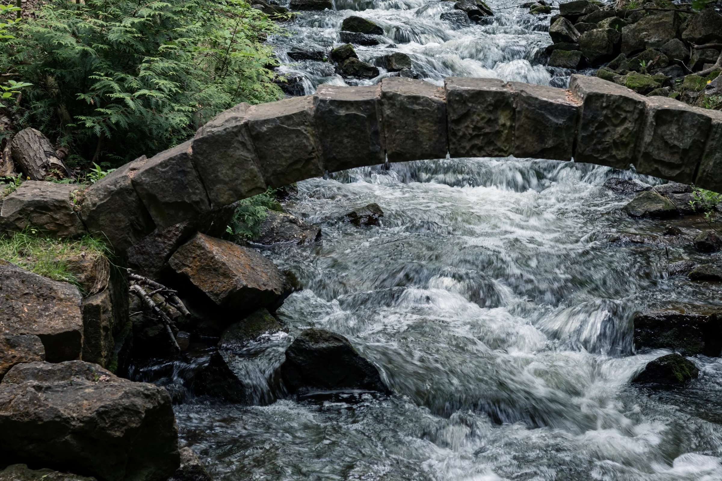 A low stone footbridge spans a rocky stream surrounded by green vegetation.
