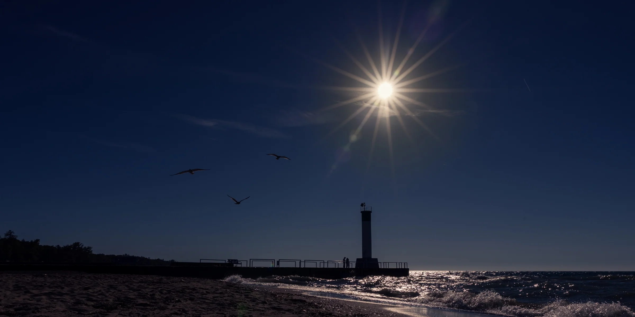 A bright sunstar shines above a silhouetted pier and beacon as birds cross the sky.
