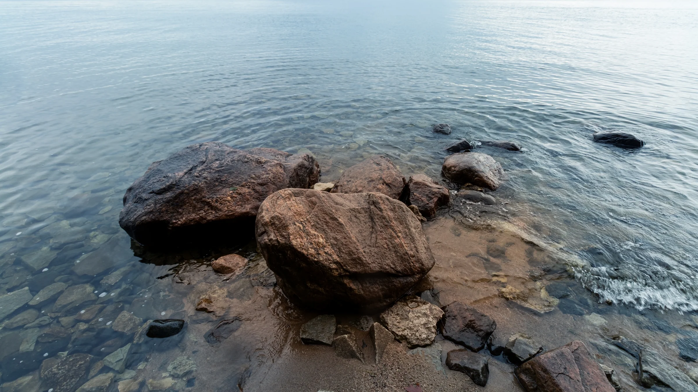 Warm brown rocks emerge from shallow, clear water along a quiet shore.