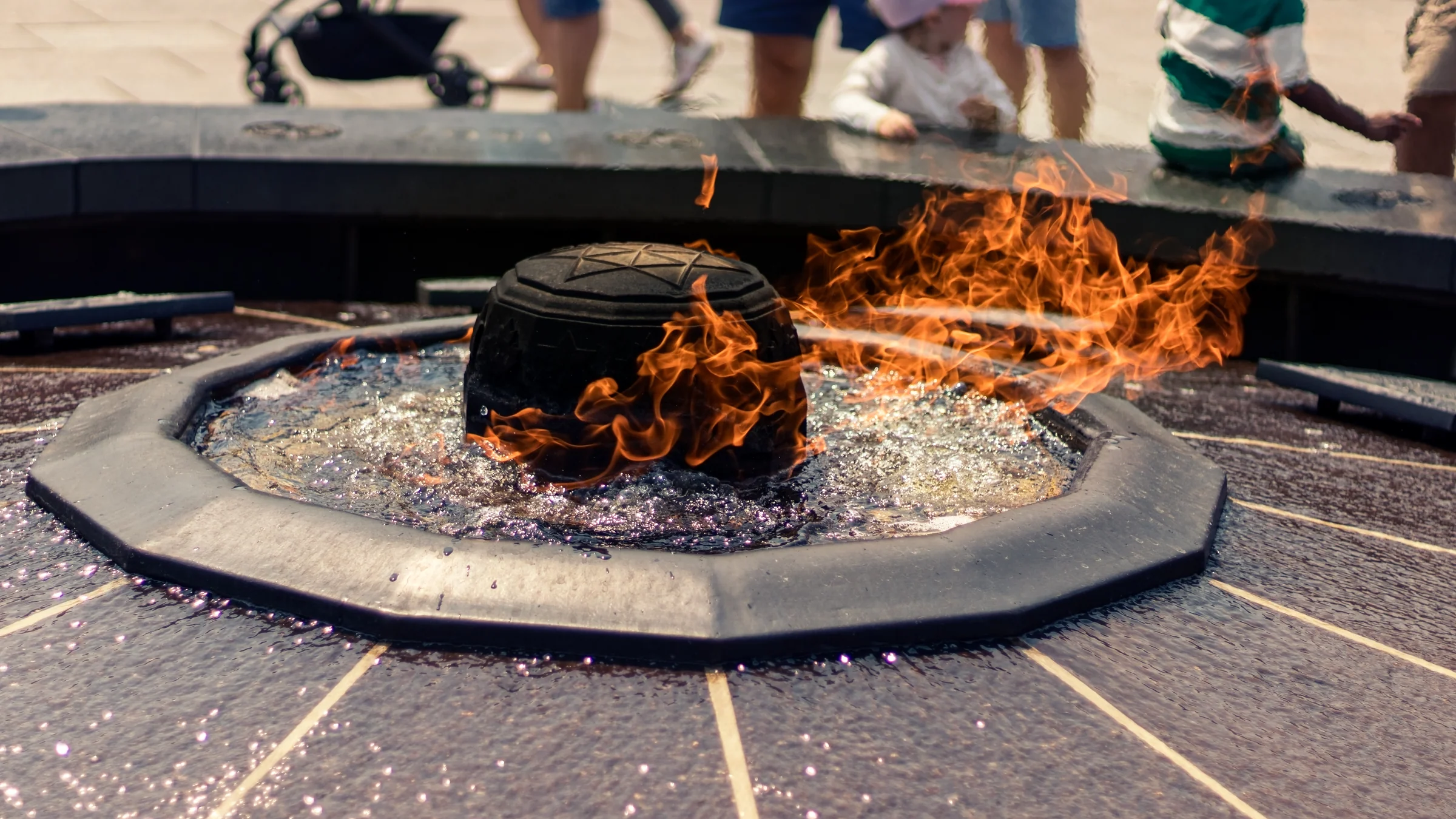 Orange flames rise from a circular outdoor fire feature as visitors gather beyond.