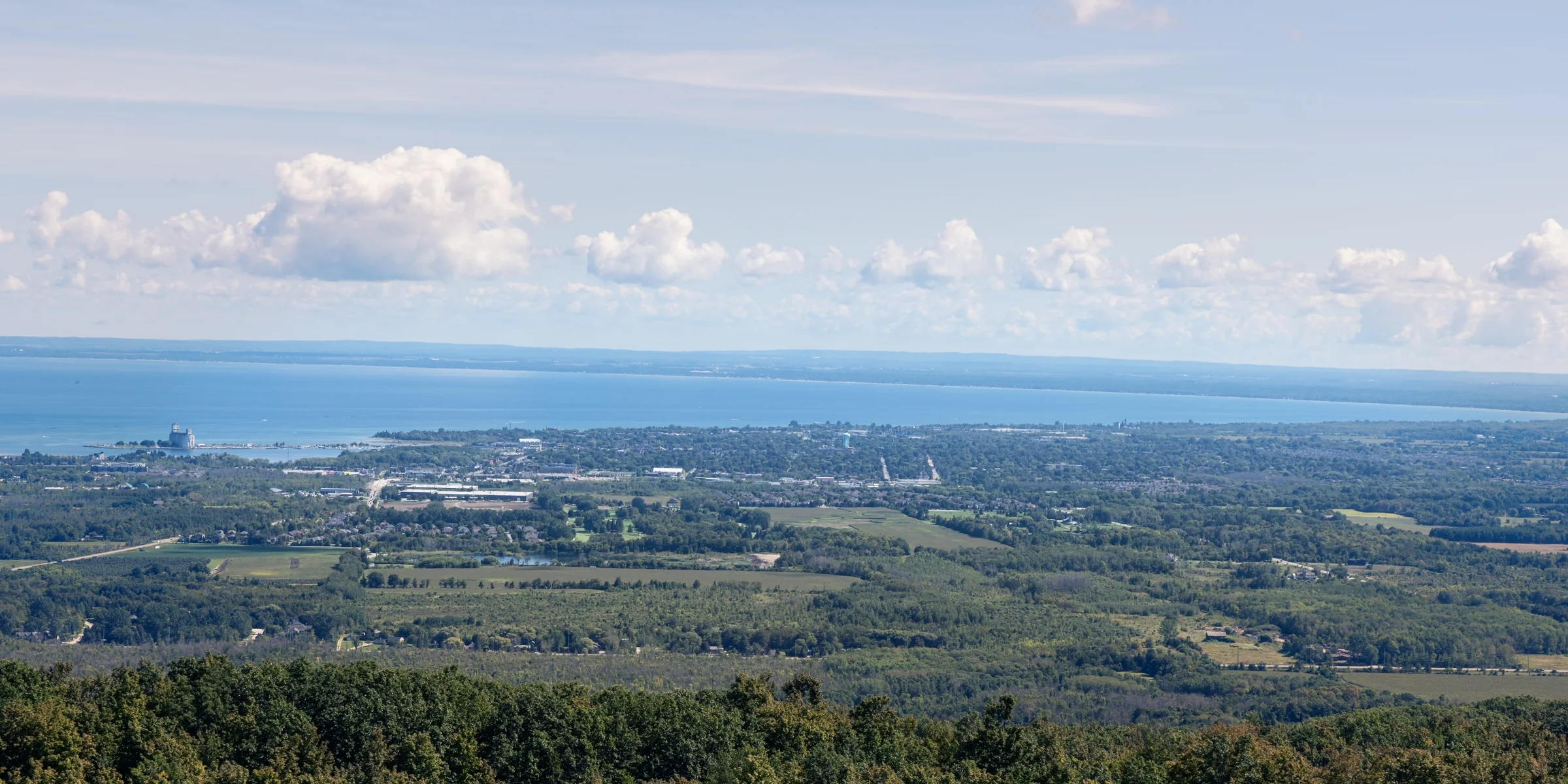 Patchwork fields and scattered settlements stretch toward distant blue water beneath white clouds.