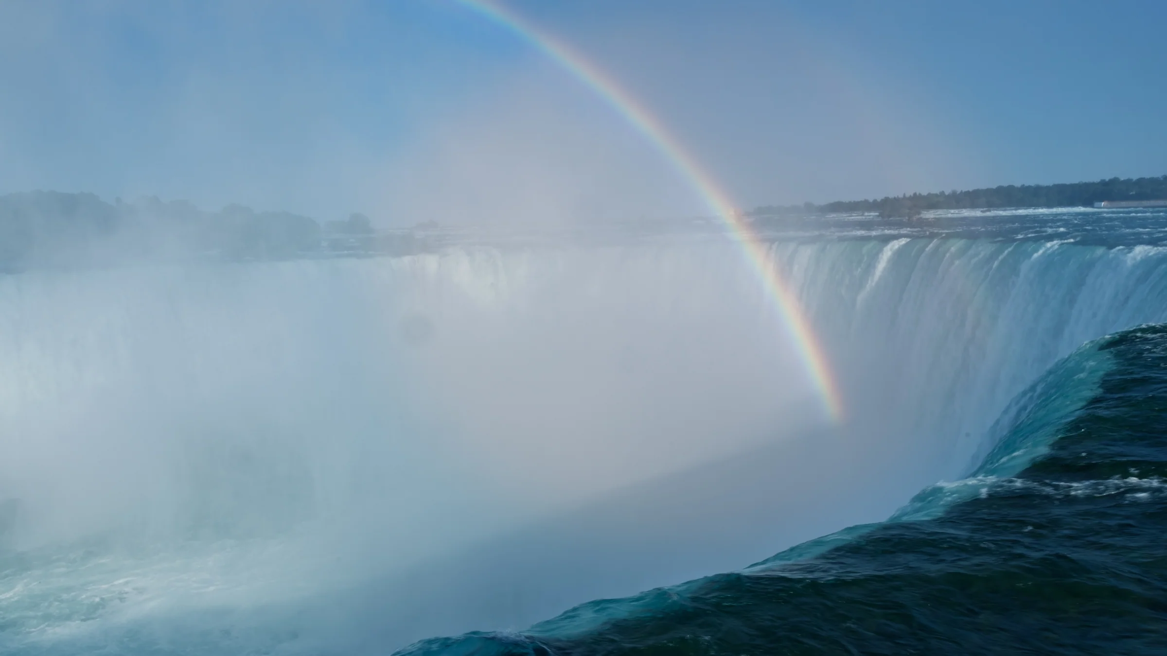 A rainbow fades into the mist beside a sweeping curtain of falling water.