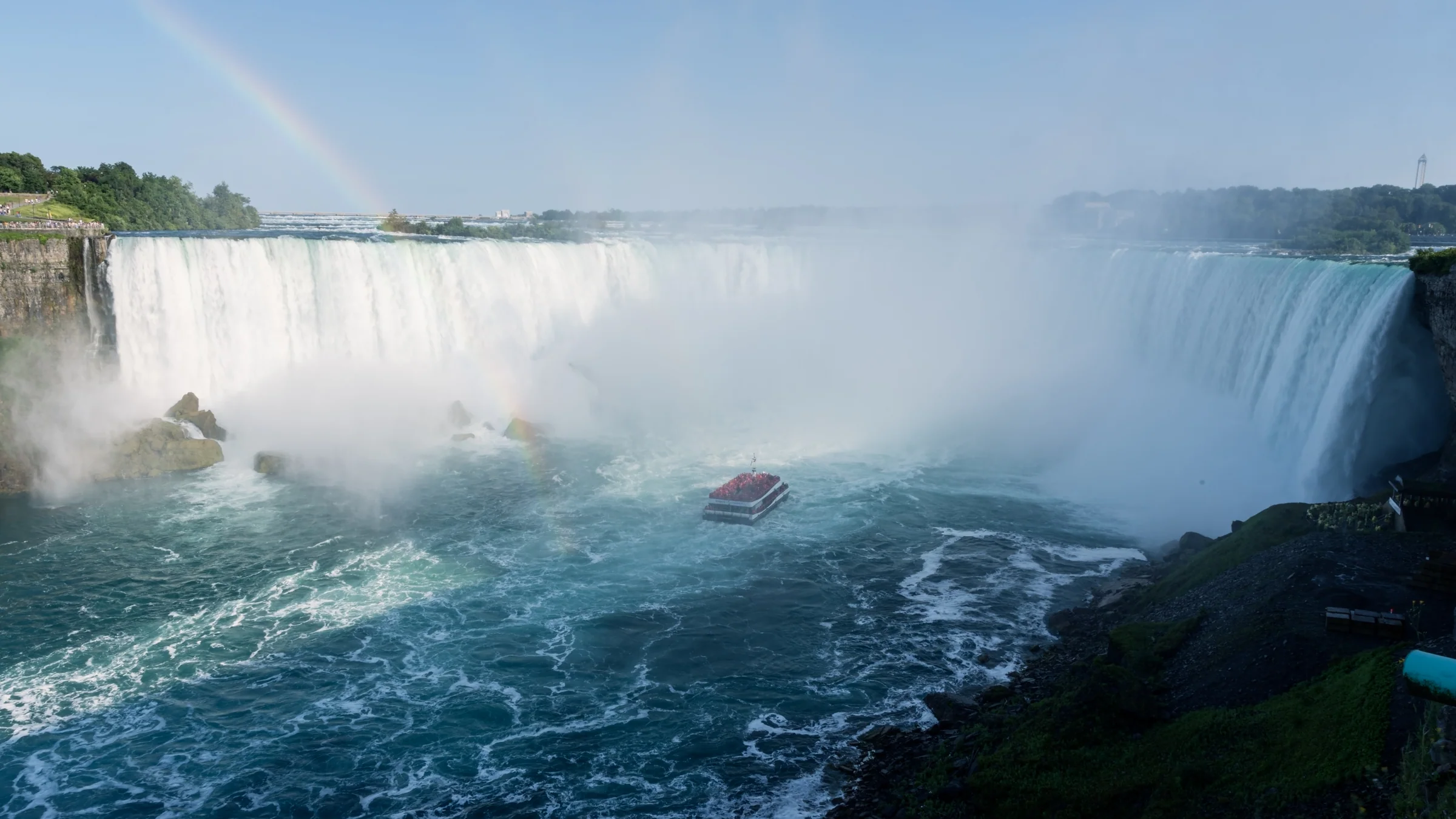 A faint rainbow rises through the spray above powerful waterfalls and dark blue water.