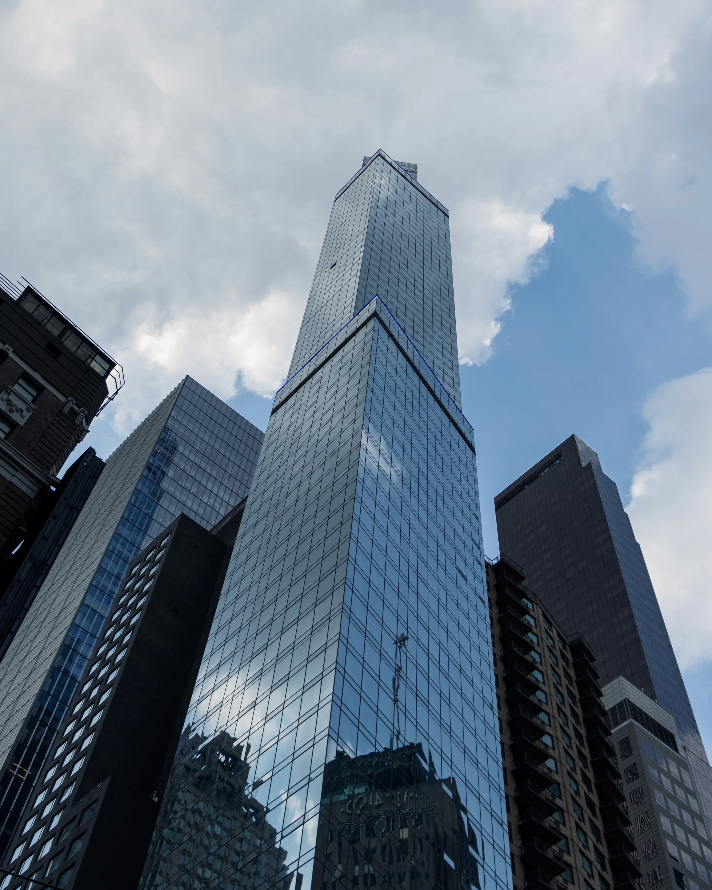 A tall glass skyscraper rises into a partly cloudy sky between neighbouring towers.