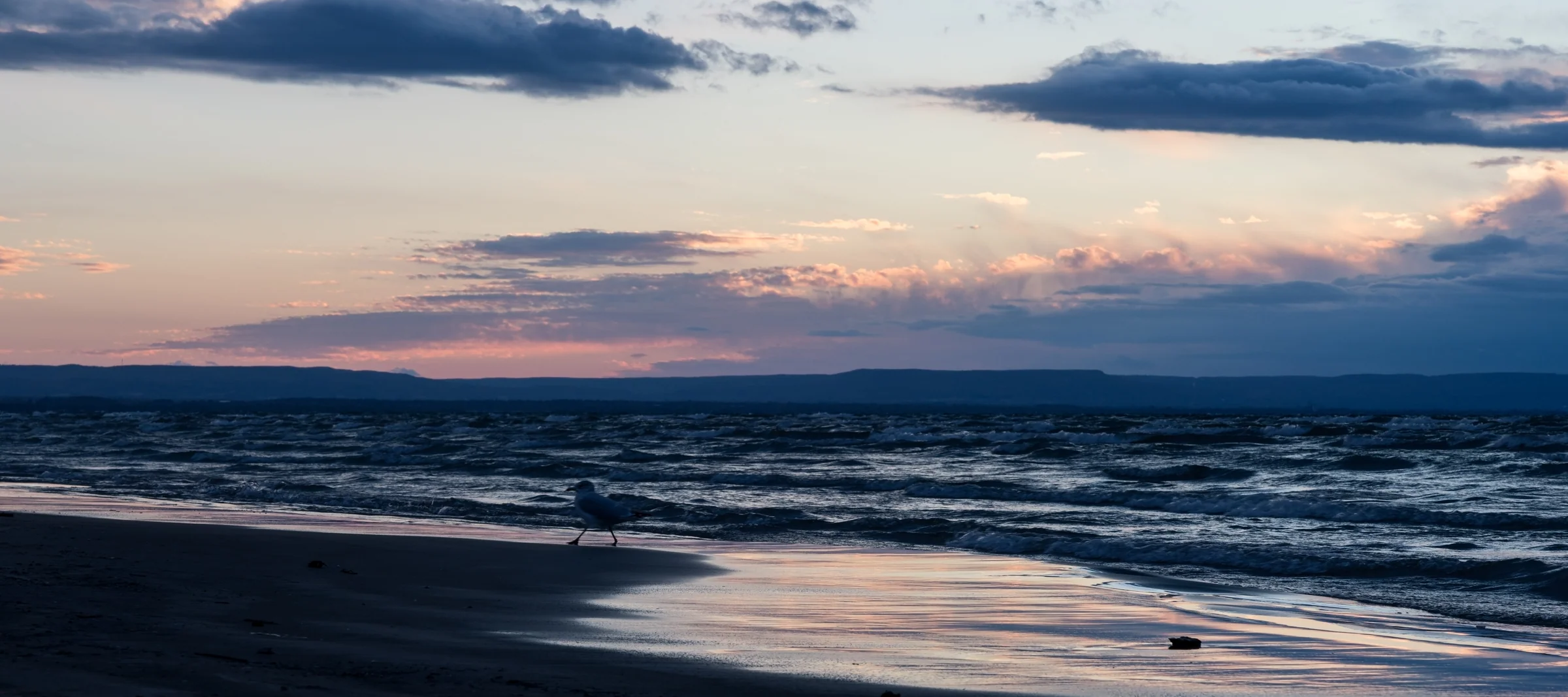 Gentle waves reflect pastel sunset colours beneath long blue-grey clouds.