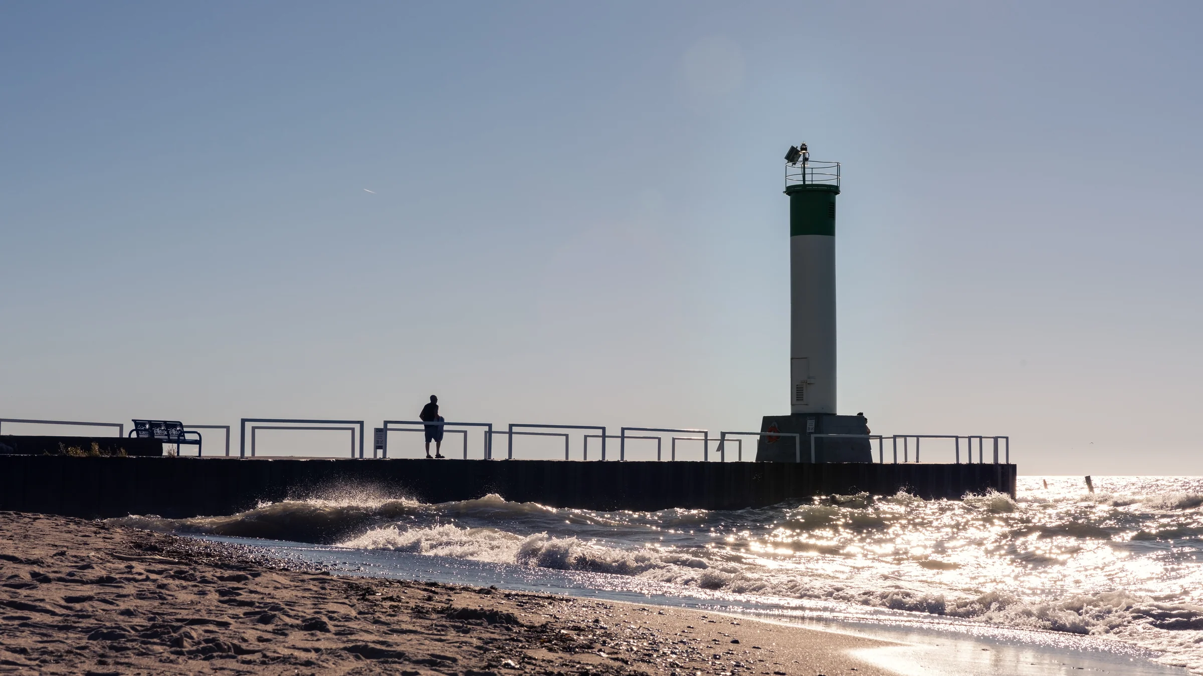 A tall green-and-white beacon stands at the end of a pier above choppy sunlit water.
