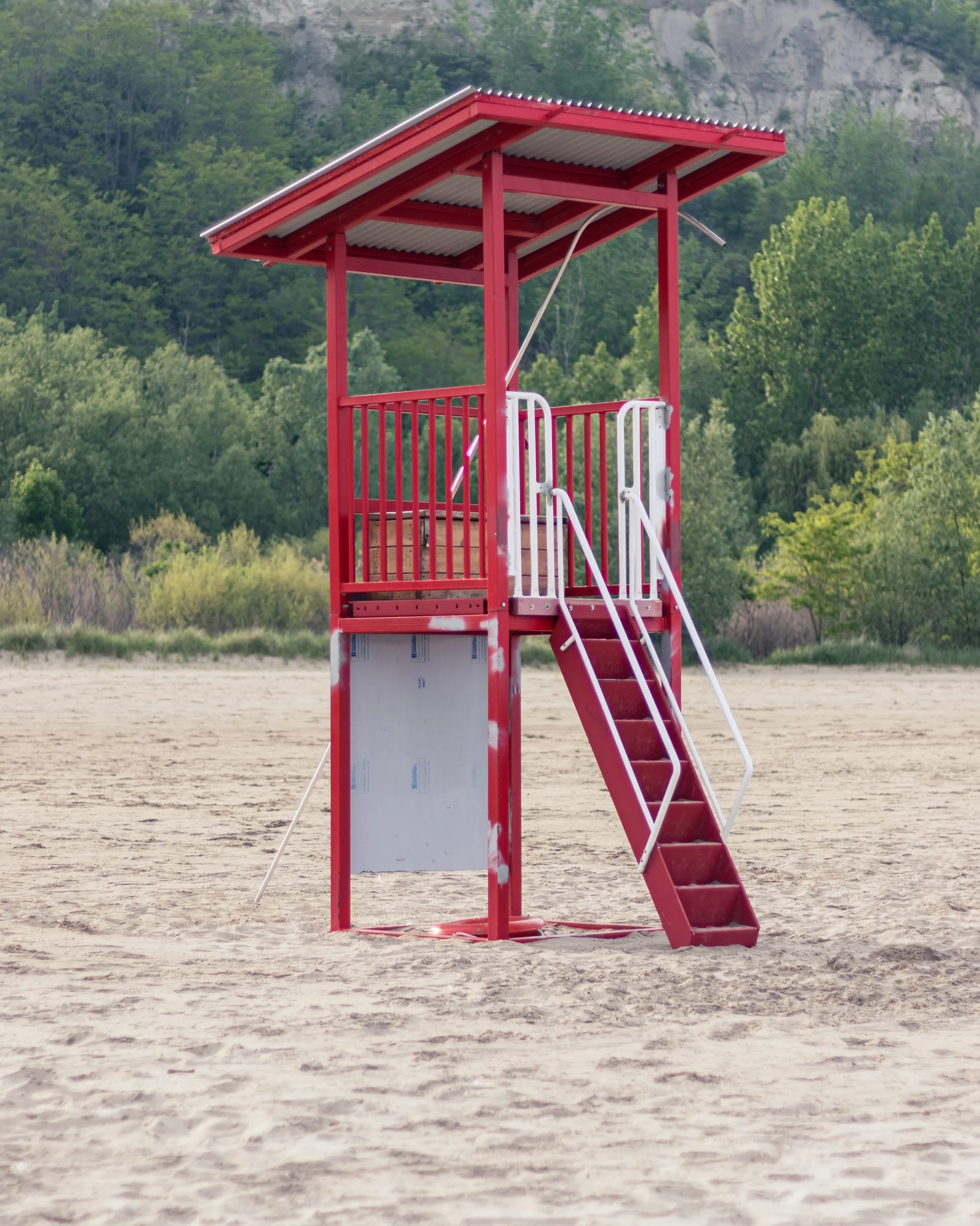 A bright red lifeguard tower stands on an empty sandy beach beside a wooded slope.