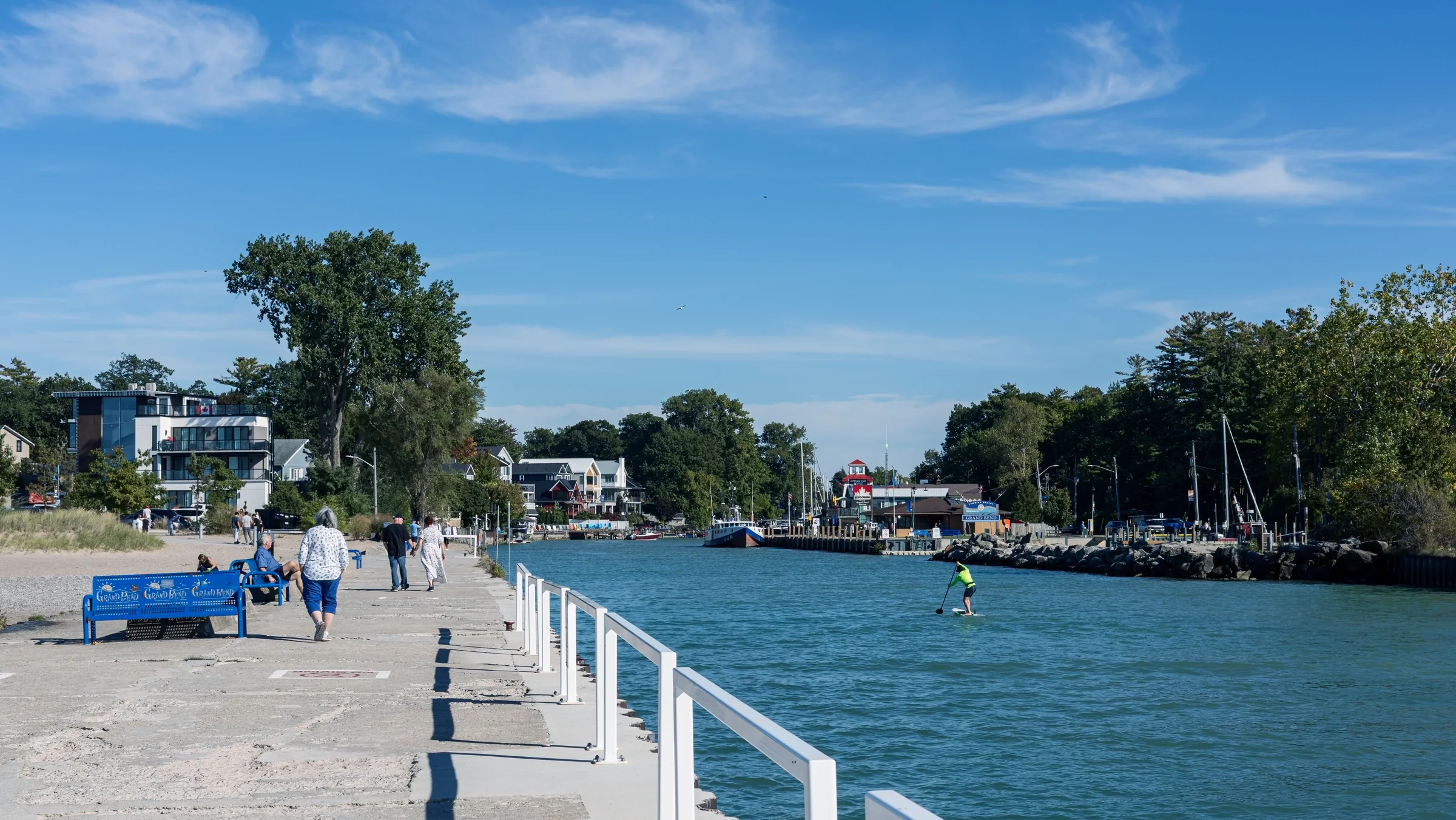 A waterside walkway follows a calm harbour lined with buildings, trees, and moored boats.