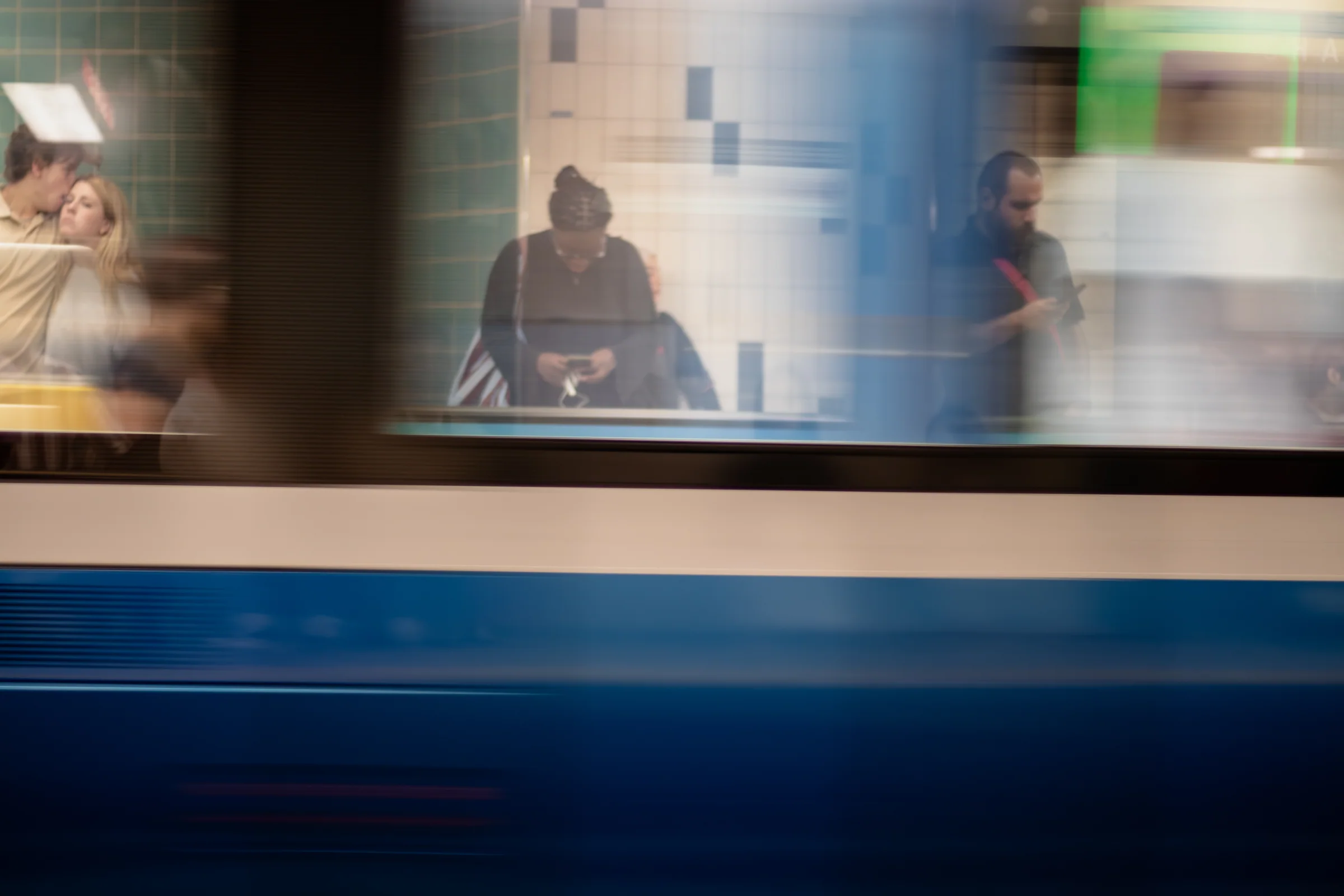 People waiting on a tiled station platform, seen through the motion blur of a passing blue and white train.