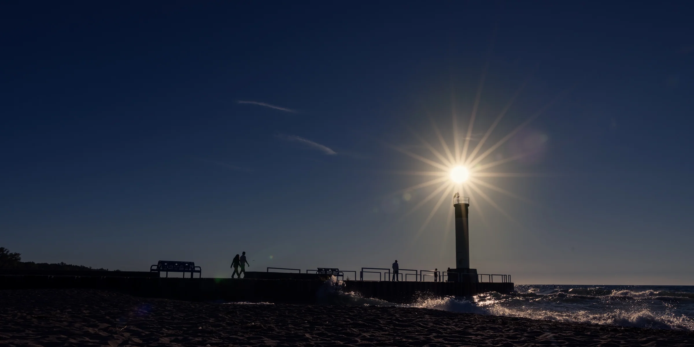 The sun forms a starburst just above a silhouetted beacon beside the water.