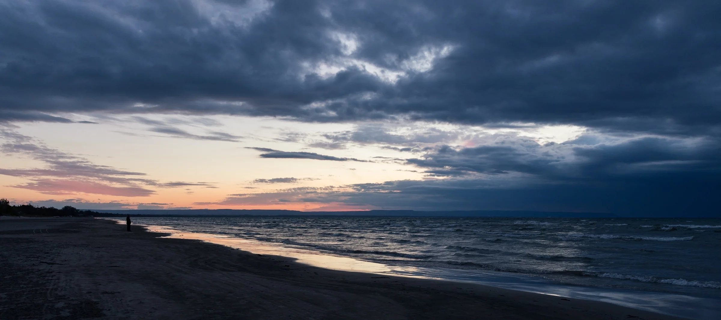 A narrow band of sunset light glows beneath heavy clouds along a dark shoreline.