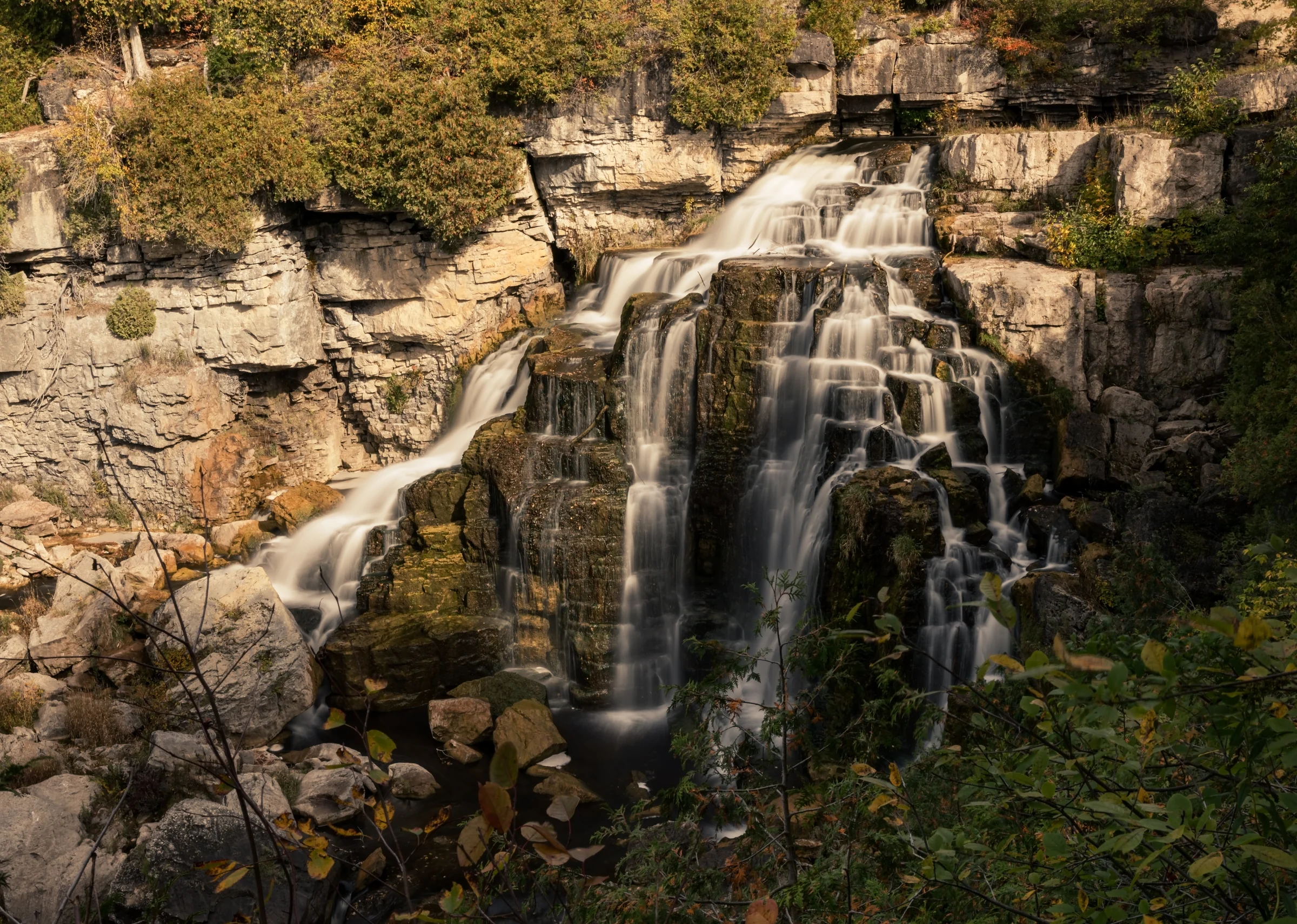 A broad cascade descends across layered pale rock surrounded by gold and green trees.